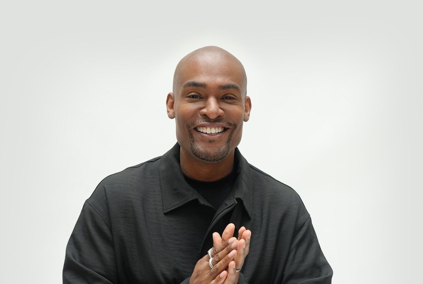 Paul C Brunson smiling towards the camera in front of a white backdrop, wearing a black tshirt and jacket